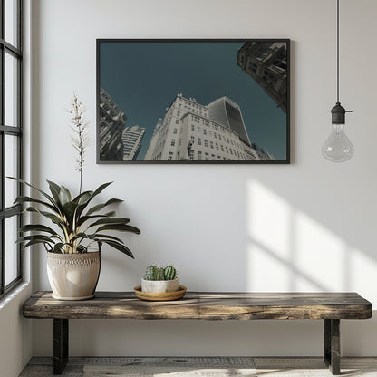 A framed wall art print depicting an upward-looking view of city skyscrapers under a dark teal sky, displayed on a white wall above a rustic wooden bench with a potted plant and a hanging light bulb in a modern room setting.
