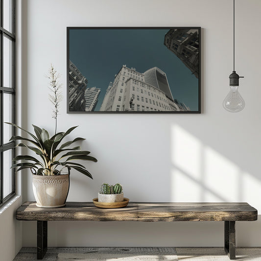 A framed wall art print depicting an upward-looking view of city skyscrapers under a dark teal sky, displayed on a white wall above a rustic wooden bench with a potted plant and a hanging light bulb in a modern room setting.