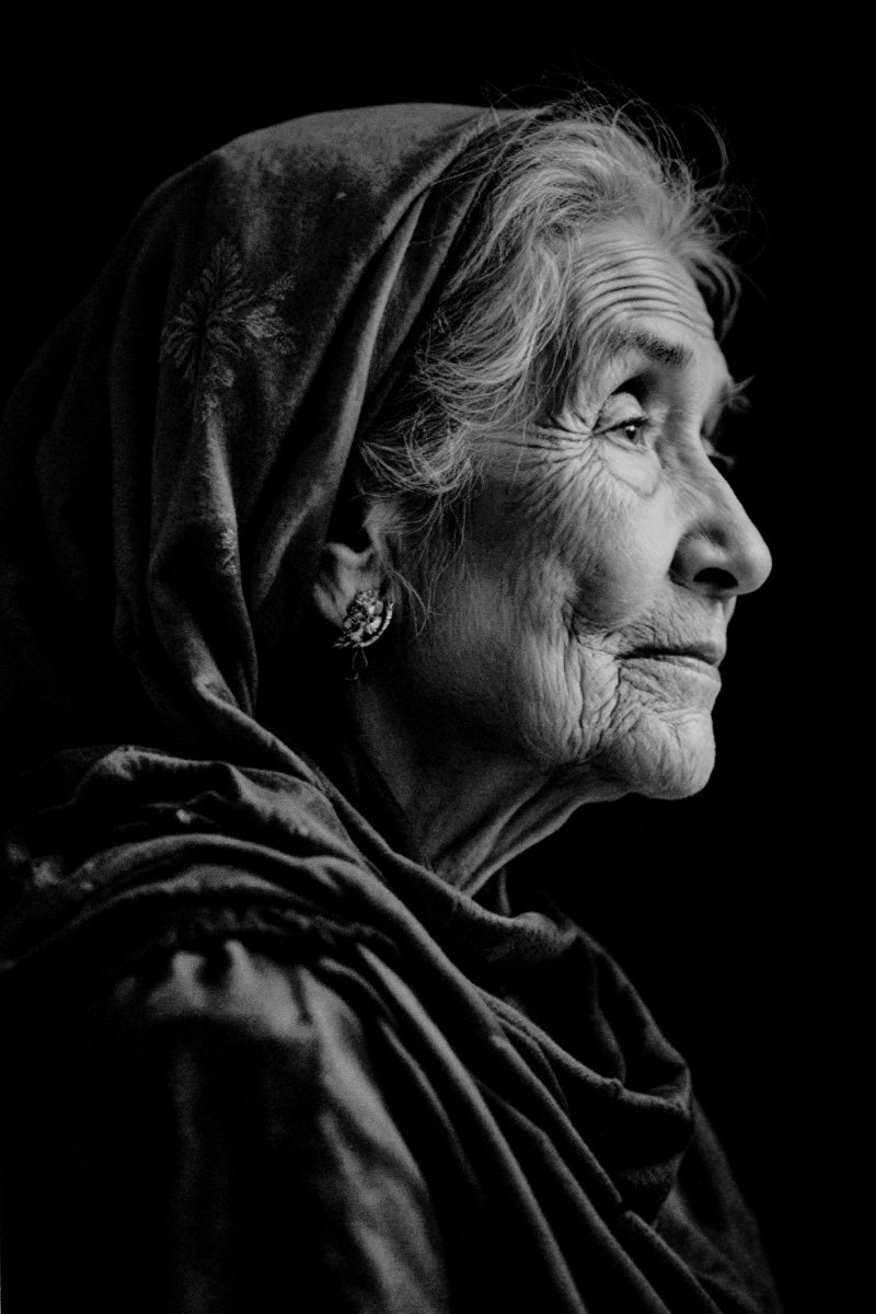 Black and white portrait of an elderly woman in profile wearing a dark headscarf, shown against a solid black background.