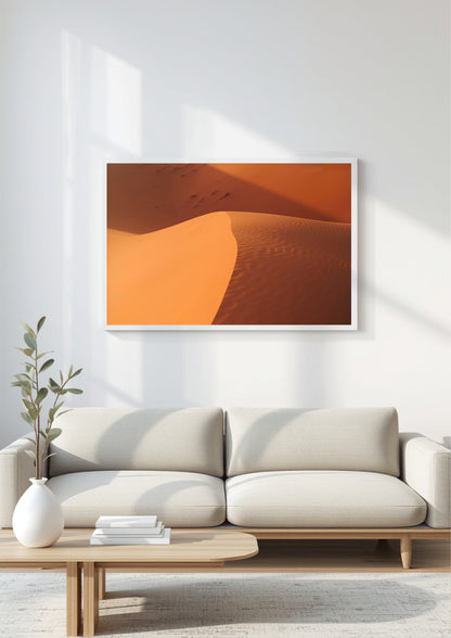 A framed wall art print showing warm orange and tan sand dunes, displayed above a light beige sofa in a bright, modern living room.
