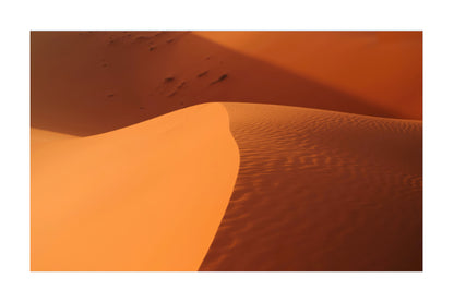 A square art print depicting warm orange and brown sand dunes with textured ripples and soft shadows, centered on a white background.