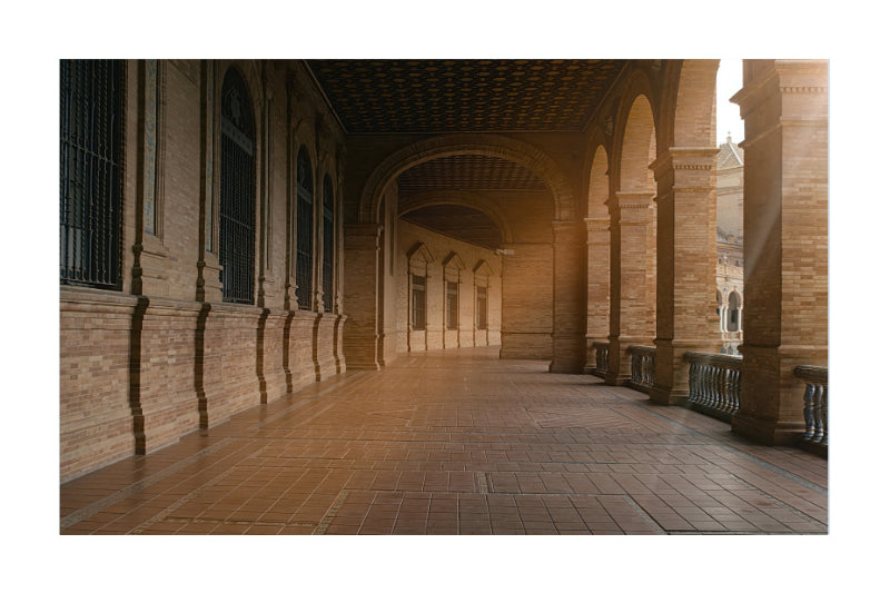 A scenic photograph of a historic covered walkway with arches and columns, featuring warm brown brick walls and tiled floors under soft sunlight.