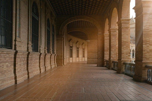 A long covered walkway with repeating arches and columns, made of warm tan stone and brick, with tiled flooring and sunlight streaming in from an open side.