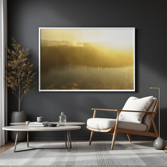 A framed landscape art print depicting a misty golden forest lake, displayed on a dark gray wall in a modern living room setting with a wooden armchair, round coffee table, and a potted plant.
