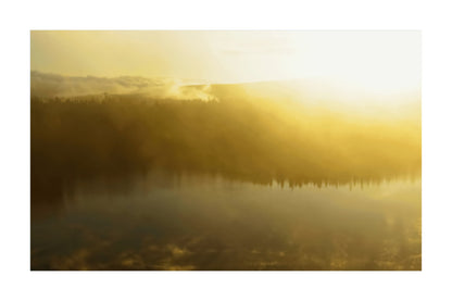 A misty golden landscape scene with a calm lake reflecting a dark treeline and mountains under bright low sunlight.