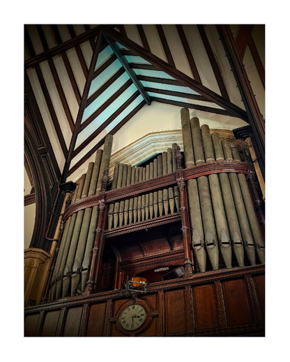 A low-angle photograph of a large historic pipe organ inside a church, framed by a white border. The organ features numerous dark metal pipes, ornate dark wood casing, and a round clock below, set against a high wooden-beamed ceiling with a light blue stained glass window.