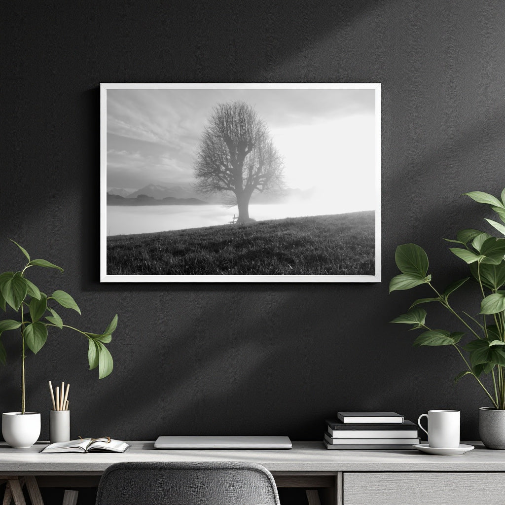 A black and white landscape art print of a solitary tree on a hill by a lake, displayed in a white frame on a dark gray wall above a modern desk with plants and books.