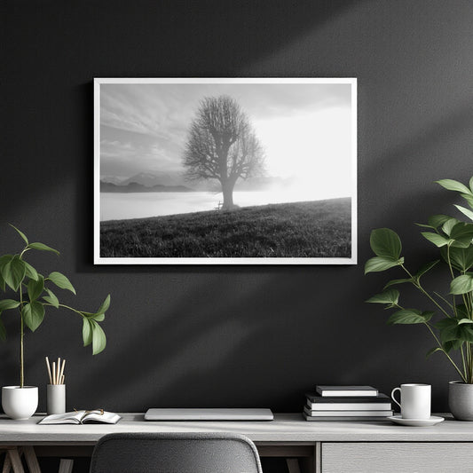 A black and white landscape art print of a solitary tree on a hill by a lake, displayed in a white frame on a dark gray wall above a modern desk with plants and books.