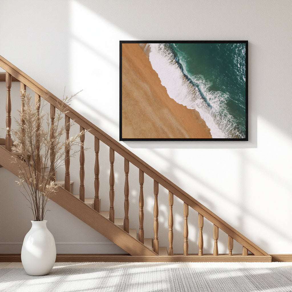 A framed square wall art print showing an aerial beach scene with turquoise ocean waves and golden sand, hung on a white wall above a wooden staircase in a bright interior.