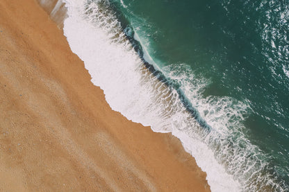 Aerial view of a sandy beach meeting the ocean, with white waves washing onto the shore and turquoise water on the right.