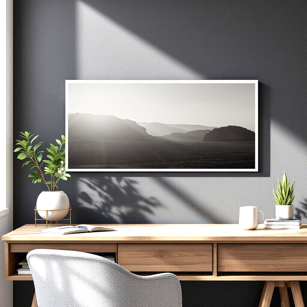 A framed black and white landscape art print hangs on a dark gray wall above a wooden desk with plants, a mug, and a chair.