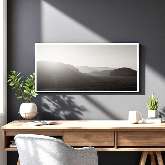 A framed black and white landscape art print hangs on a dark gray wall above a wooden desk with plants, a mug, and a chair.