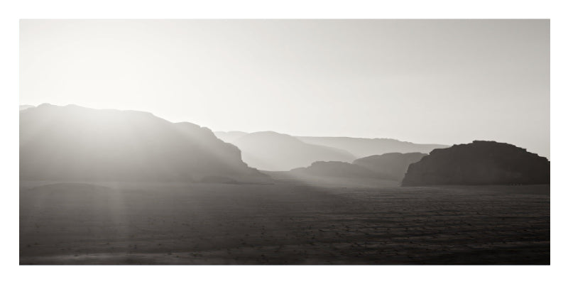 A black and white landscape photograph featuring layered mountain ranges under a bright sun, with hazy light rays filtering over a flat plain.