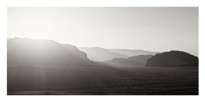 A black and white landscape photograph featuring layered mountain ranges under a bright sun, with hazy light rays filtering over a flat plain.