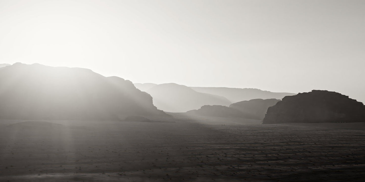 Black and white landscape photograph of layered mountain ranges under a bright sun, with hazy atmospheric light and a dark foreground plain.