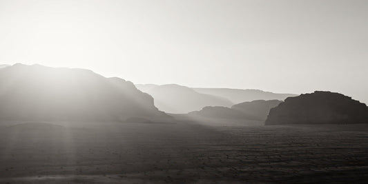 Black and white landscape photograph of layered mountain ranges under a bright sun, with hazy atmospheric light and a dark foreground plain.