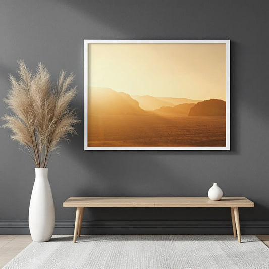 A framed landscape art print featuring a warm golden desert sunset hangs on a dark gray wall above a light wood bench with a white vase and pampas grass.