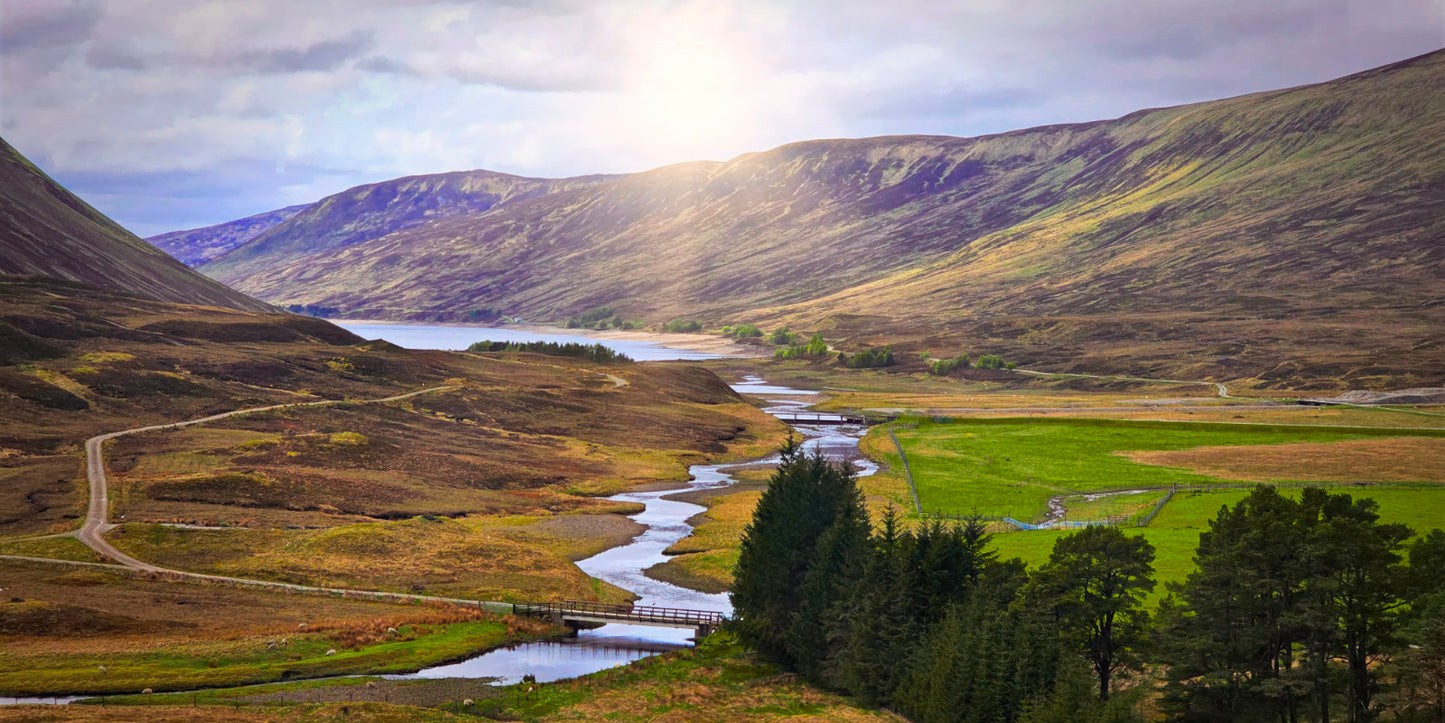 A scenic landscape photograph of a winding river flowing through a green valley between rolling hills and mountains under a dramatic cloudy sky with sunlight breaking through.