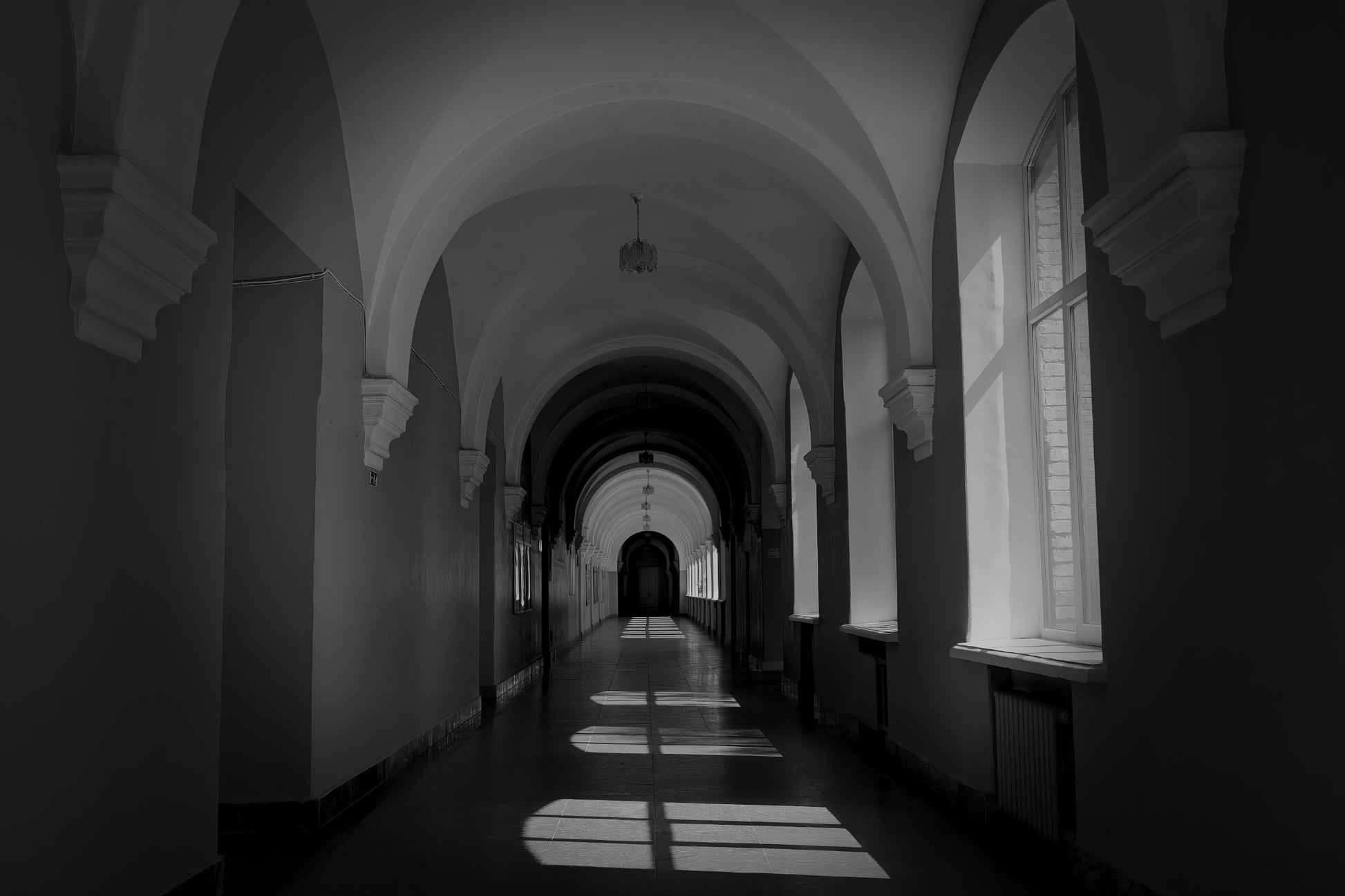 Black and white photograph of a long arched hallway with tall windows casting rectangular light patterns on the floor.
