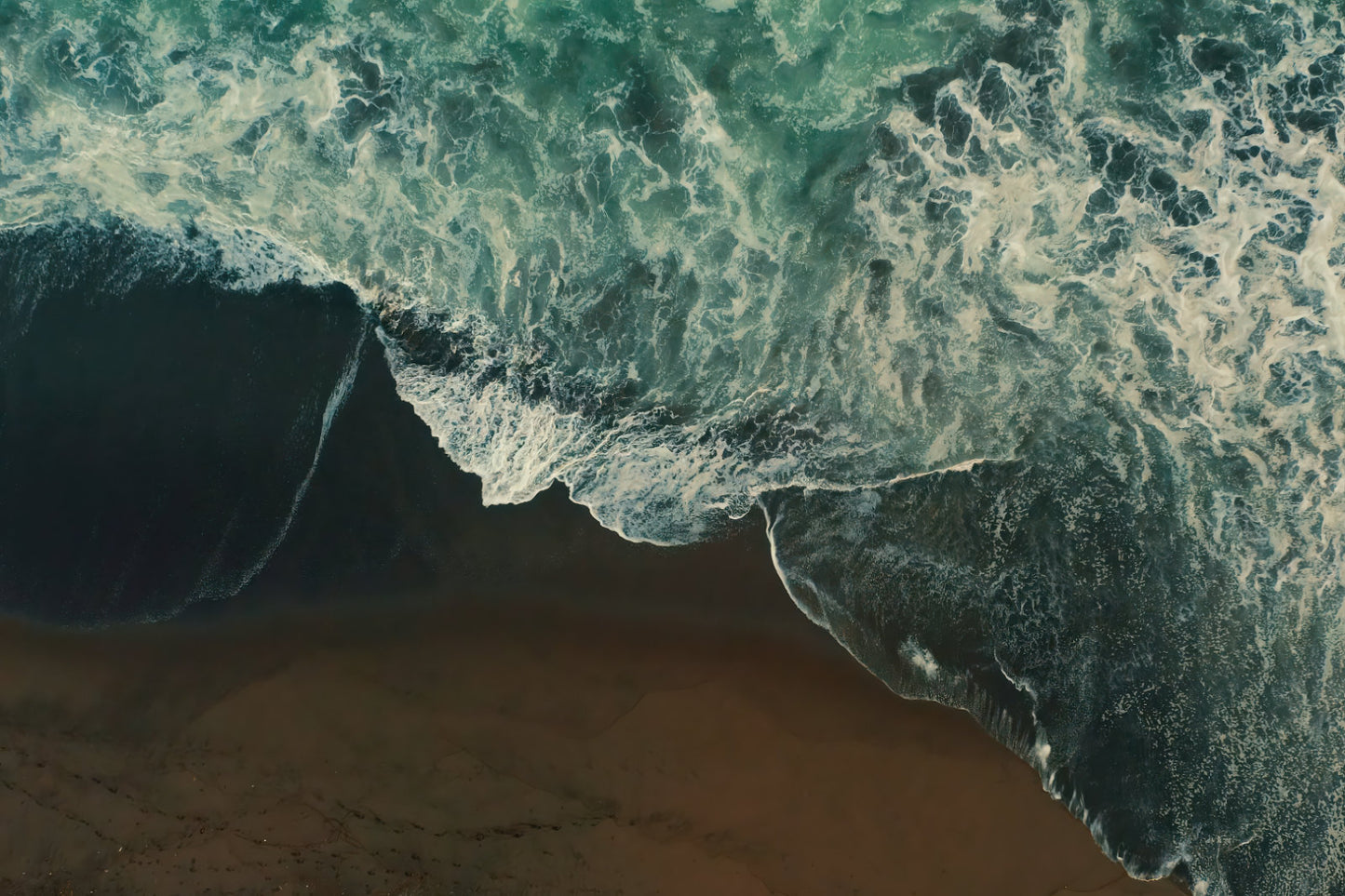Aerial view of ocean waves with turquoise and dark green water crashing onto a sandy beach.
