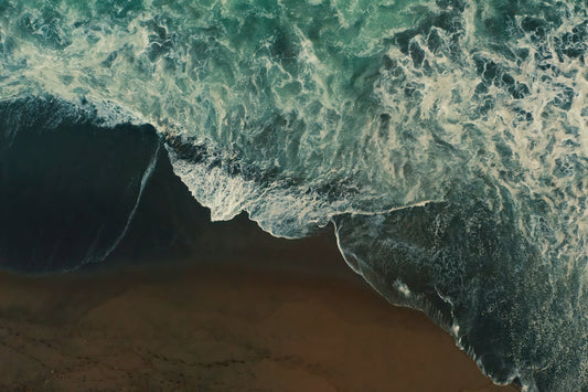 Aerial view of ocean waves with turquoise and dark green water crashing onto a sandy beach.