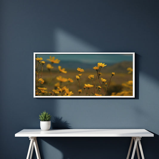 A framed horizontal art print depicting yellow wildflowers in a field under a soft blue sky, displayed on a dark blue wall above a white console table with a small potted plant.