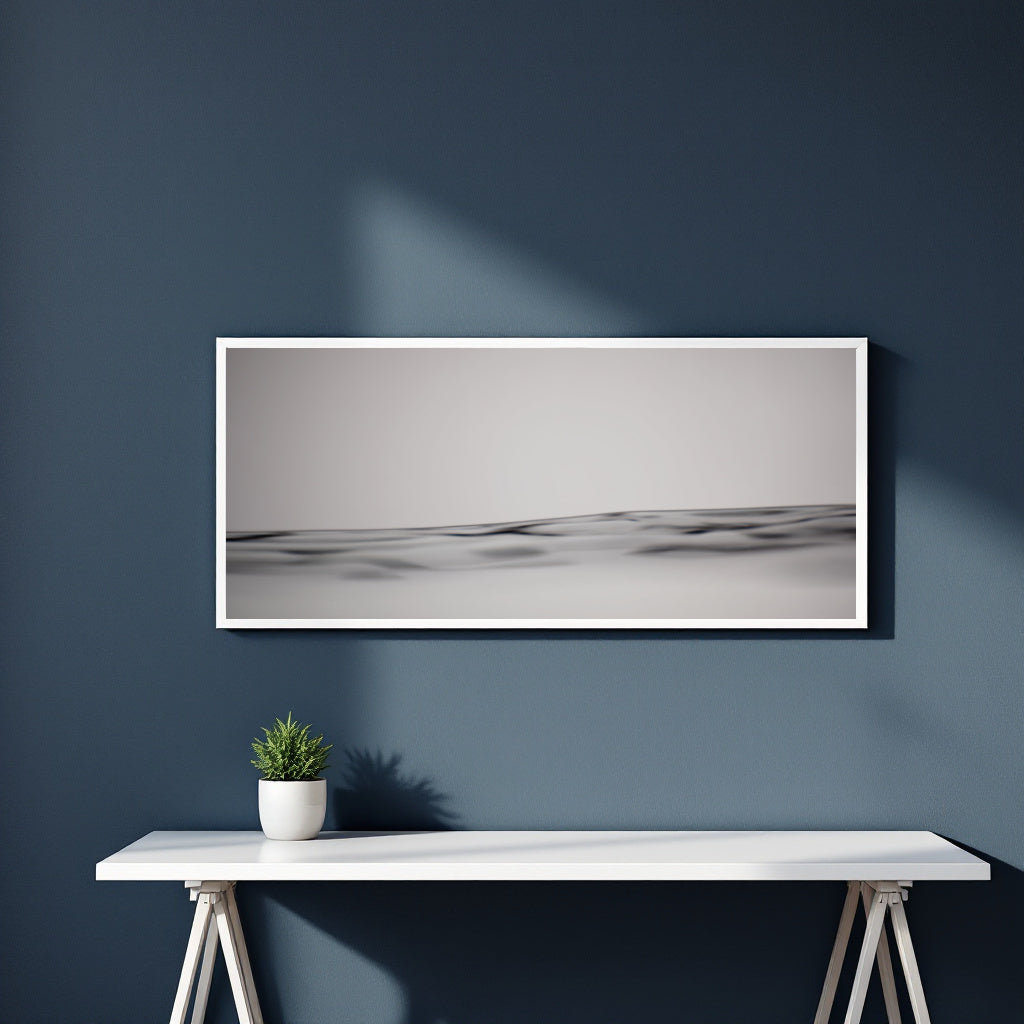 A framed horizontal black and white landscape photograph of rolling dunes, displayed on a dark blue wall above a white console table with a small potted plant.