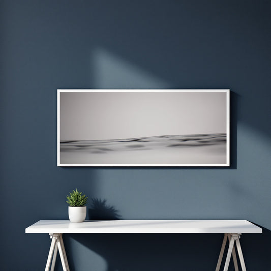 A framed horizontal black and white landscape photograph of rolling dunes, displayed on a dark blue wall above a white console table with a small potted plant.
