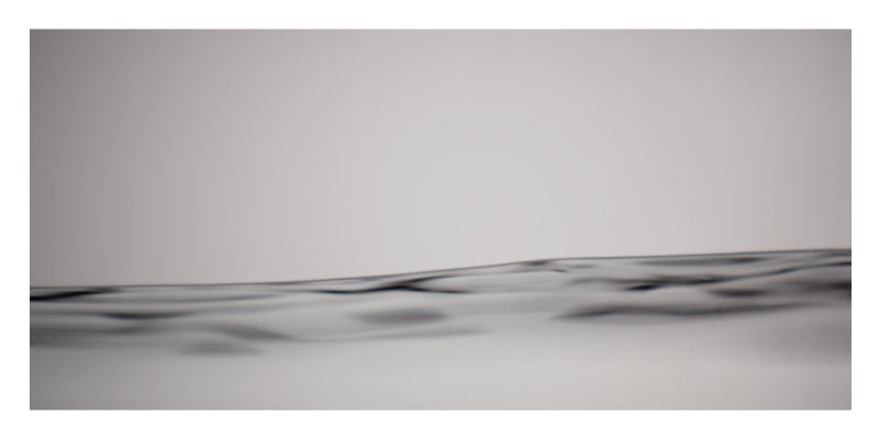 A minimalist abstract photograph of a dark, textured, undulating surface stretching horizontally across the bottom half of a square frame, set against a solid light gray background.