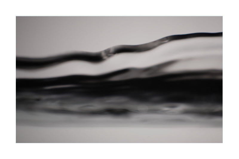 A close-up abstract photograph of layered, wavy translucent bands in black, gray, and white, framed with white borders on the left and right.