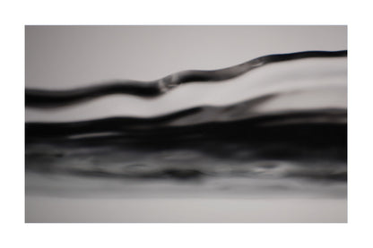 A close-up abstract photograph of layered, wavy translucent bands in black, gray, and white, framed with white borders on the left and right.