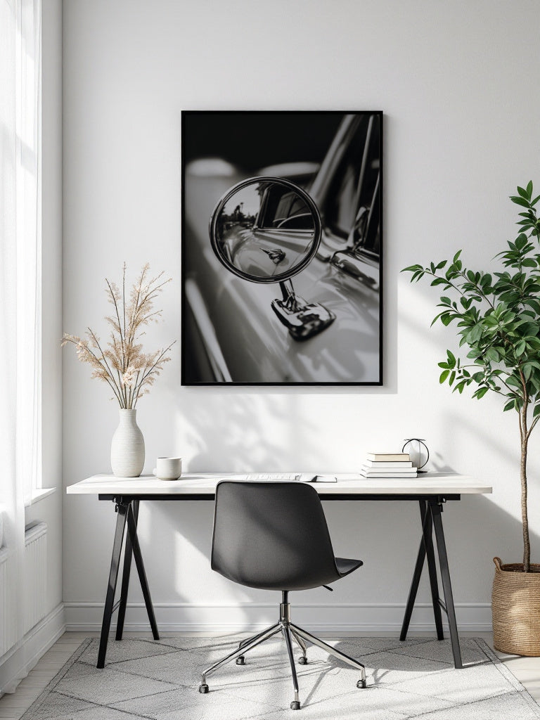 A black and white framed wall art print depicting a close-up of a vintage car side mirror, displayed in a modern home office setting above a white desk with a black chair.