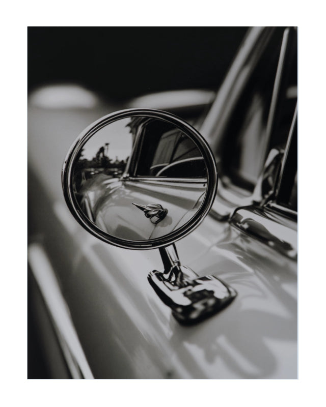 Black and white close-up photograph of a classic car side-view mirror mounted on the door, reflecting the car's chrome details and surroundings.