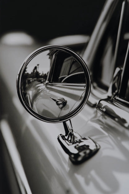 A close-up black and white photograph of a classic car's chrome side-view mirror mounted on the door.