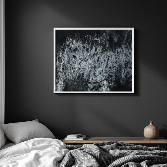 A framed black and white abstract wave art print hanging on a dark gray wall above a bed with white bedding and a wooden side table.