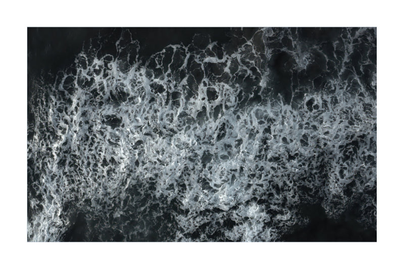 A square photographic print or poster depicting an aerial view of white ocean waves and foam against a dark water background, centered on a white border.