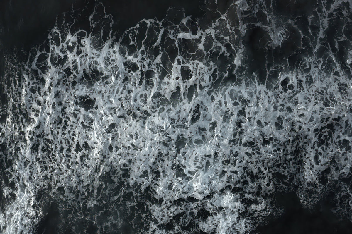 A close-up aerial view of foamy white ocean waves breaking over dark water, filling the frame.