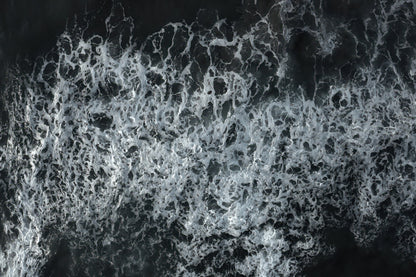 A close-up aerial view of foamy white ocean waves breaking over dark water, filling the frame.