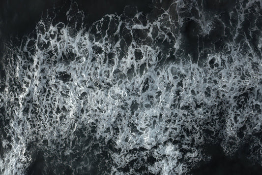 A close-up aerial view of foamy white ocean waves breaking over dark water, filling the frame.