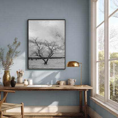 A black and white framed wall art print depicting a bare tree in a snowy landscape, hung on a light blue wall above a wooden desk in a styled room with a window, lamp, and dried flowers.
