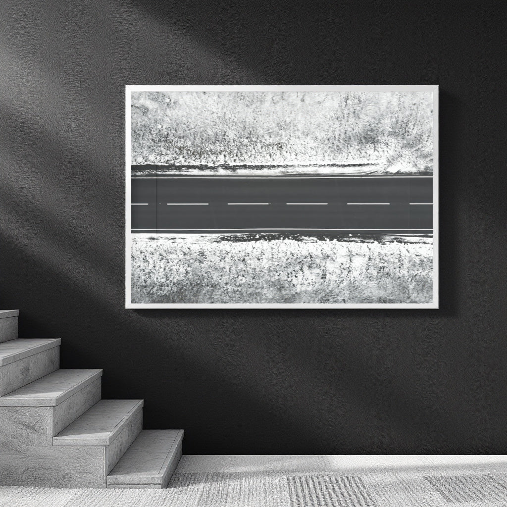 A framed black and white art print depicting an aerial view of a road cutting through textured fields, displayed on a dark wall above a staircase.