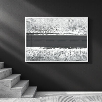 A framed black and white art print depicting an aerial view of a road cutting through textured fields, displayed on a dark wall above a staircase.