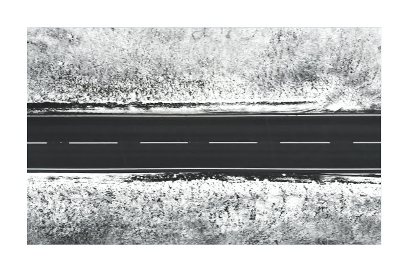 A black and white aerial photograph of a straight road with dashed white lines, bordered by dense, textured vegetation.