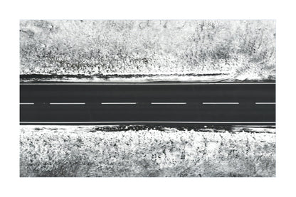 A black and white aerial photograph of a straight road with dashed white lines, bordered by dense, textured vegetation.