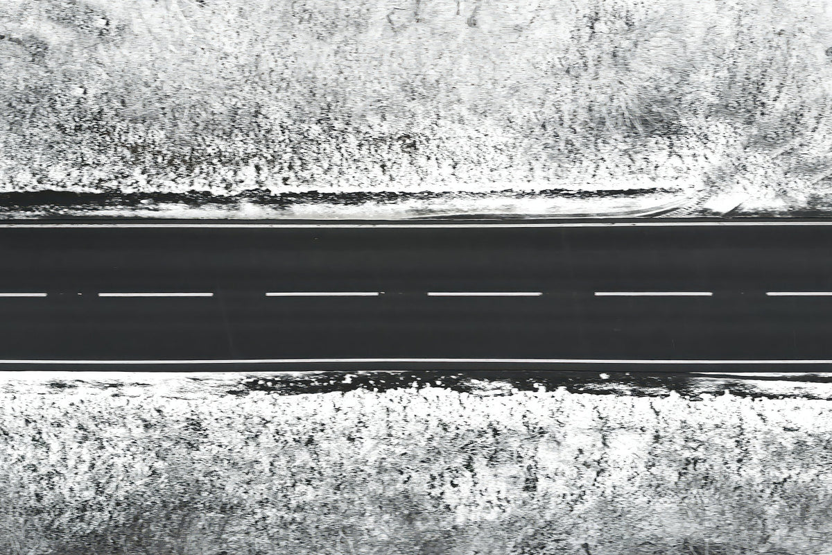 A black aerial view of a straight two-lane road with dashed white lines, bordered by snowy, textured ground on both sides.