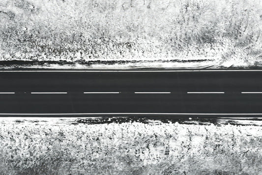A black aerial view of a straight two-lane road with dashed white lines, bordered by snowy, textured ground on both sides.
