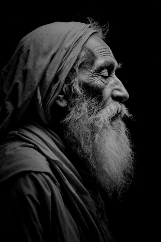 Black and white portrait of an elderly man with a long beard and head covering, shown in profile against a dark background.