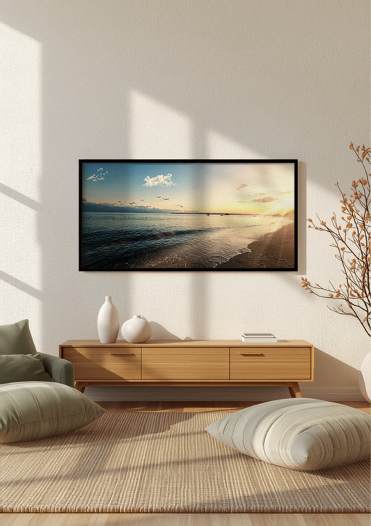 A framed wall art print depicting a beach sunset over calm water, displayed in a modern living room setting above a light wood console table with decorative vases and cushions.